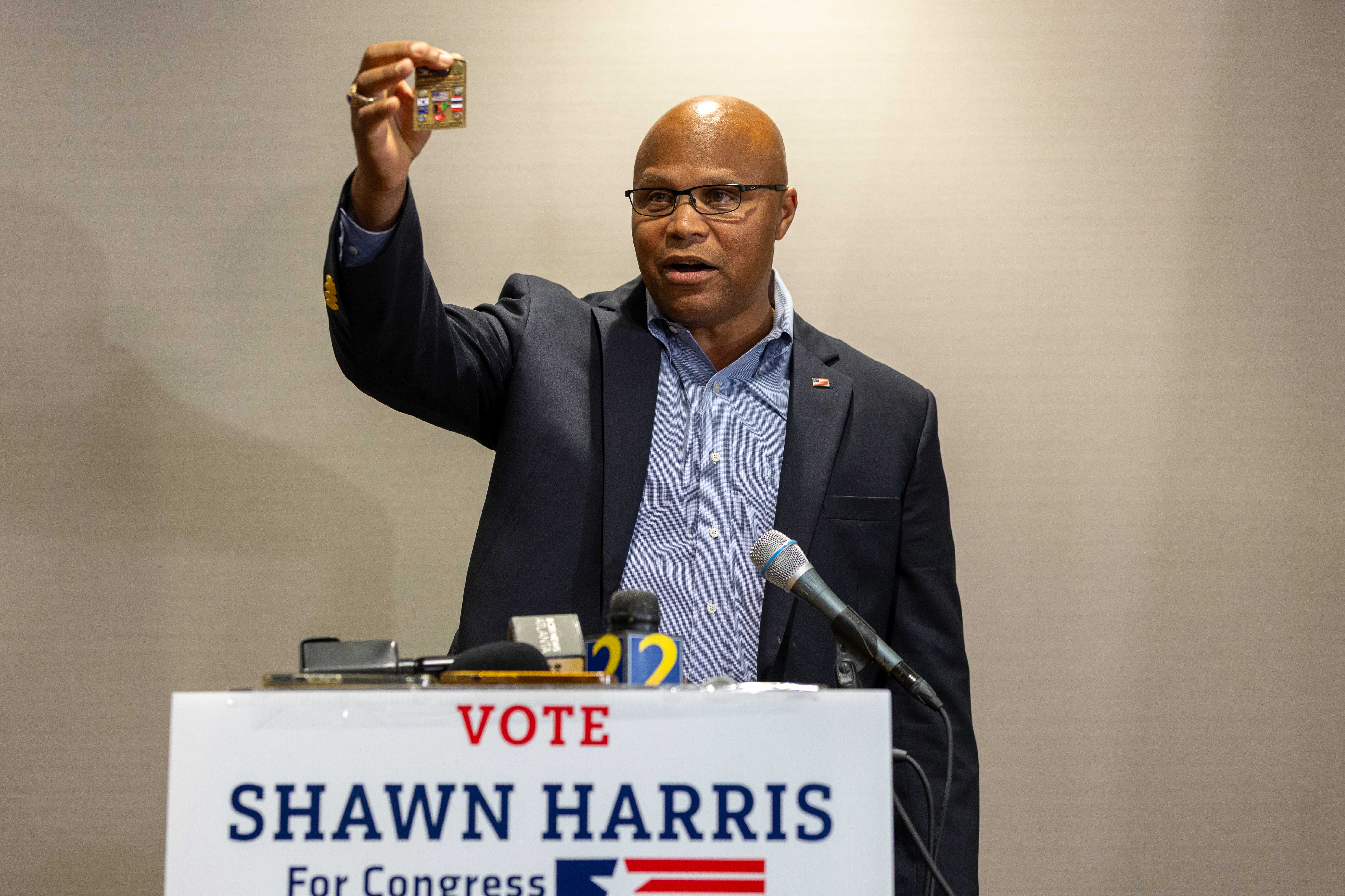 A man in a suit stands behind a podium with microphones, holding up an item. The podium features a "Vote Shawn Harris For Congress" sign. The atmosphere is serious.