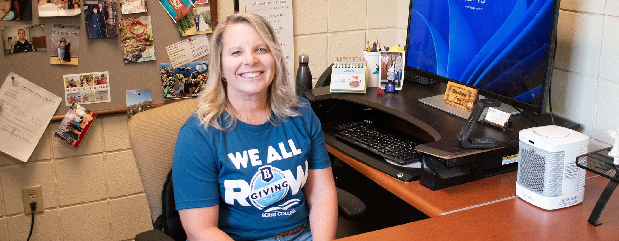 Smiling woman sitting at a desk in an office, wearing a blue t-shirt that says 'WE ALL RUN'. A computer monitor displays the time, and there are various items and photos on the wall behind her.