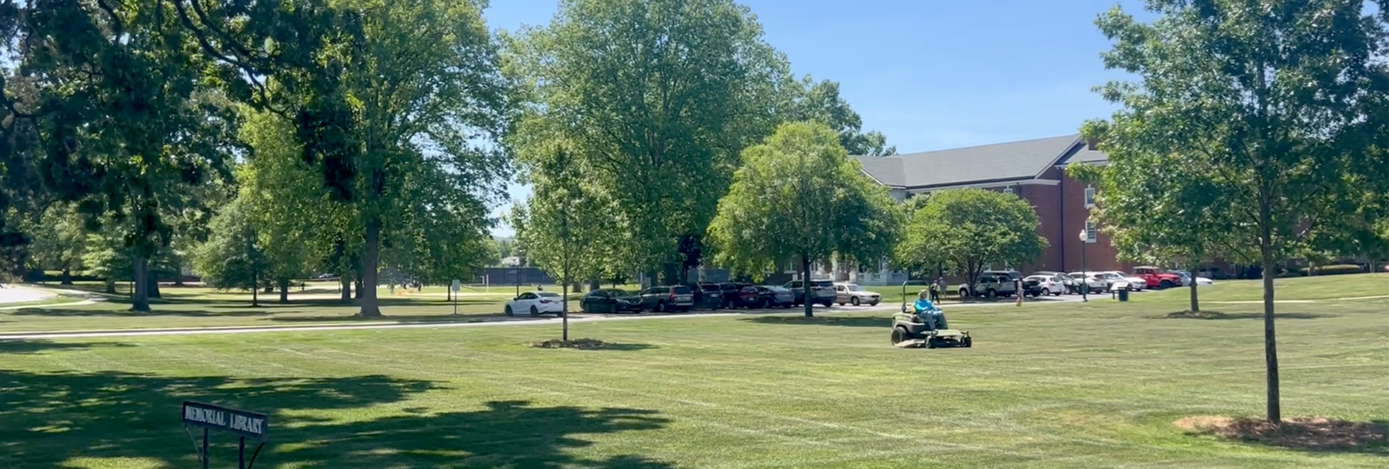 A Berry College grounds crew member mows a campus lawn bordered by trees and academic buildings.
