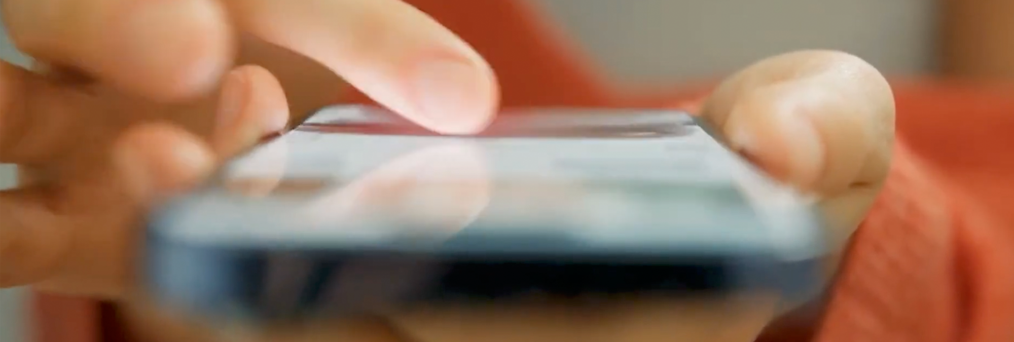 Close-up view of hands holding and scrolling on a smartphone. The image is intentionally blurred, with a finger touching the screen and warm lighting in the background, illustrating mobile device and social media use.