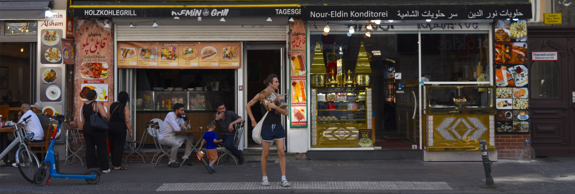 Street-level view of storefronts along a busy Berlin sidewalk. A restaurant with outdoor seating occupies the left side of the image, with menu photos displayed beside the entrance and several people seated or standing near tables. A blue rental scooter and a bicycle are parked at the curb. In the center foreground, a pedestrian carrying shopping bags stands near the street. On the right, a pastry shop with signage in both German and Arabic features illuminated gold-colored display cases and large food photographs around the entrance. Additional pedestrians, shop signs, and urban details line the commercial streetscape.