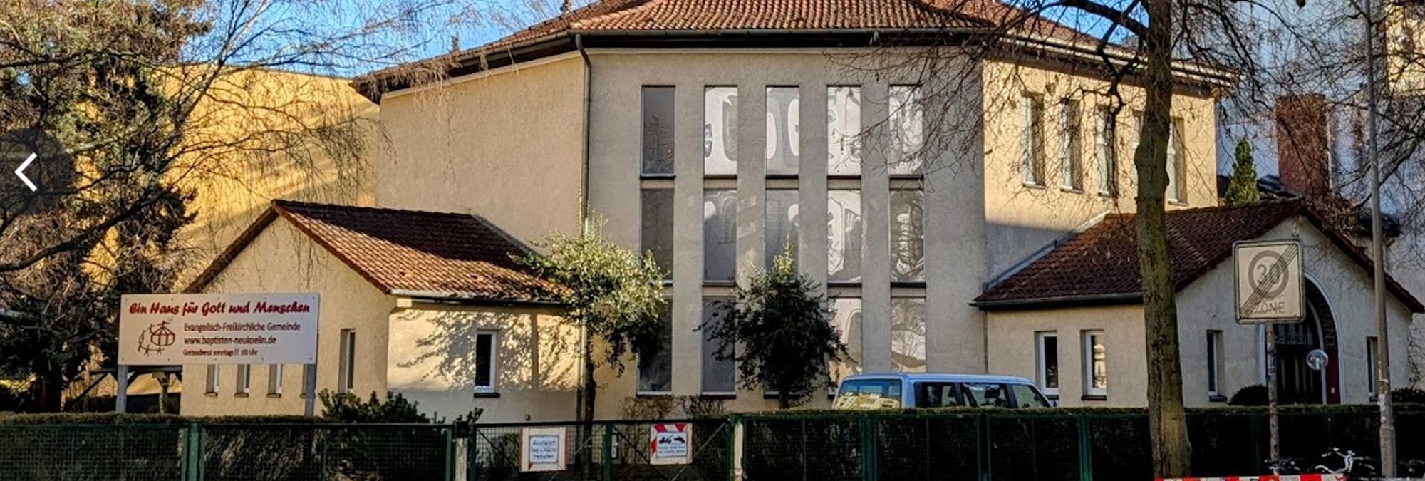 Exterior view of a church building on a tree-lined street in Berlin. The light-colored structure features a red-tiled roof, tall vertical windows, and two lower wings extending from the main building. A fenced entrance sits in the foreground with a parked van visible inside the property. A sign near the street reads, “Ein Haus für Gott und Menschen” and identifies the congregation as the Evangelisch-Freikirchliche Gemeinde (Baptist church) Neukölln. Bare tree branches frame the scene, and a traffic sign marking the end of a 30 km/h zone stands near the sidewalk. Sunlight illuminates the building and surrounding grounds on a clear day.