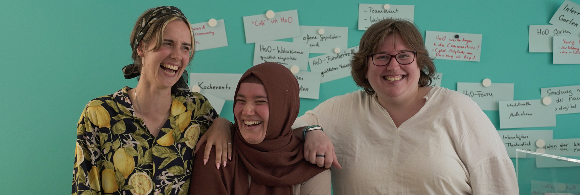 Three people stand together in front of a turquoise wall covered with handwritten notes pinned to display boards. One person wears a patterned shirt with a lemon design and a headscarf, another wears a brown head covering and light-colored clothing, and a third wears glasses and a white shirt. The group stands with their arms around one another while posing for a photograph. Behind them, the notes contain handwritten ideas and project concepts in German, creating the appearance of a collaborative community or workshop space.