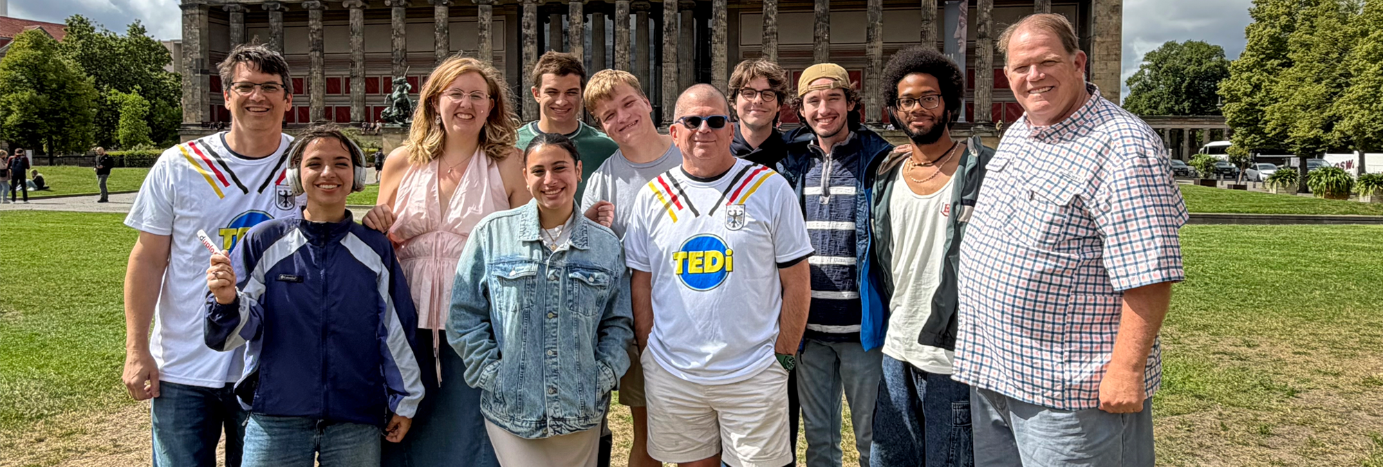 A group of ten people pose together on a grassy lawn in front of a large neoclassical museum building with tall stone columns. The group stands in two rows and wears a mix of casual clothing, including jackets, T-shirts, shorts, and jeans. Two people in the front row wear white jerseys featuring the colors of the German flag. Trees and landscaped grounds surround the museum, while partly cloudy skies provide bright daylight. The image captures a group gathering at a cultural landmark in Berlin.