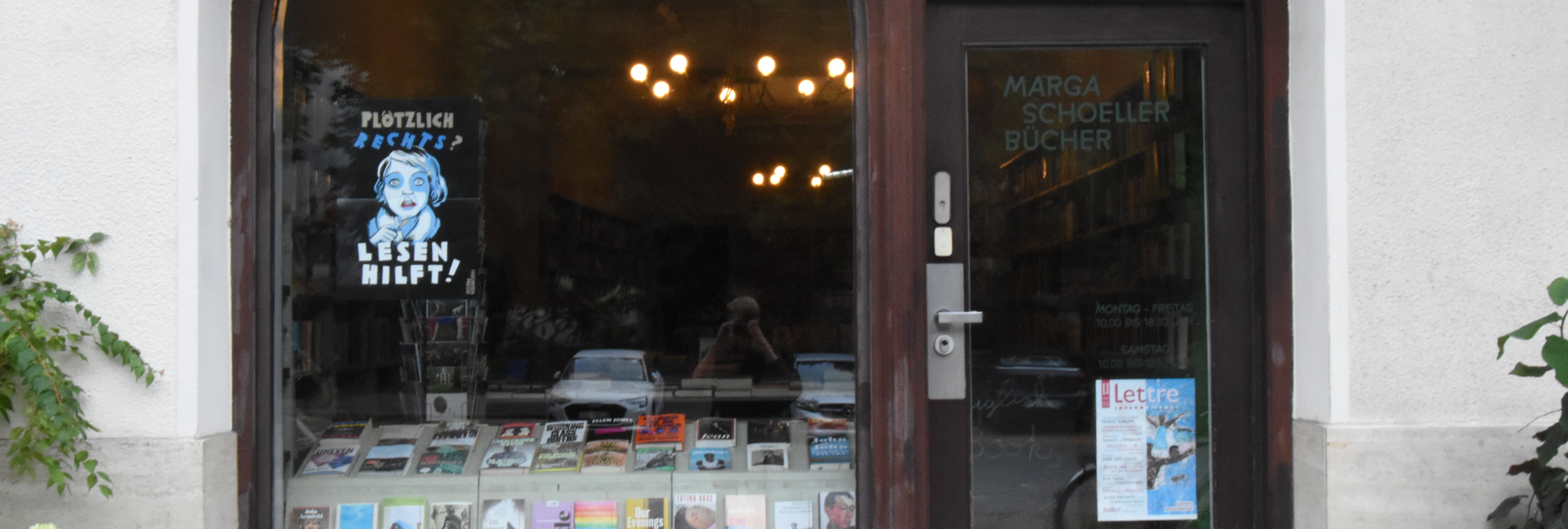 Front entrance of an independent bookstore with large arched windows and a dark wooden door. Books are displayed in the lower portion of the window, while bookshelves and warm hanging lights are visible inside the shop. Text on the door reads “Marga Schoeller Bücher,” along with the store’s opening hours. A poster in the left window features an illustrated face and the German text “Plötzlich Rechts? Lesen hilft!” (“Suddenly right-wing? Reading helps!”). Another poster advertising a literary publication is attached to the door. Reflections of parked cars and the street appear in the glass, and climbing greenery frames the storefront on both sides.