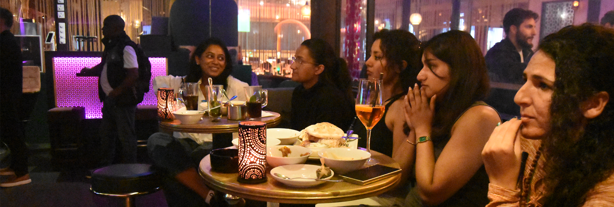 Several people sit around small tables in a dimly lit restaurant or lounge, gathered around food and drinks while watching something off-camera. Decorative candle-style lights illuminate the tables, which hold plates, bowls, glasses, and a smartphone. Purple accent lighting and warm ambient lamps create a colorful interior atmosphere. Additional patrons and seating areas are visible in the background through large windows, giving the venue a lively, social feel.