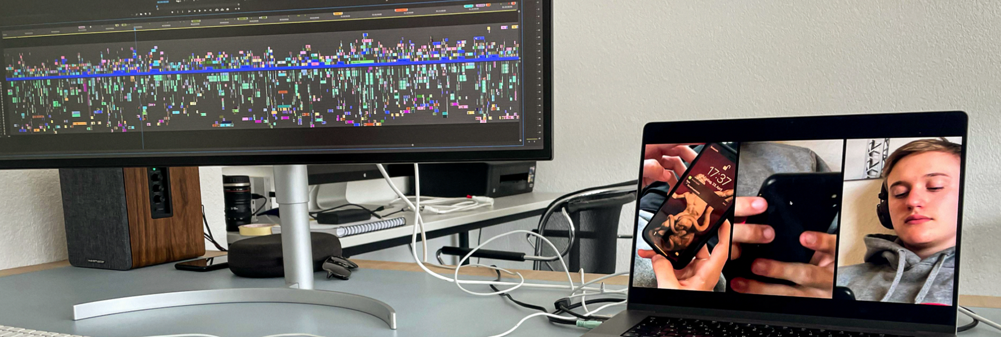 A video-editing workstation with multiple screens displays footage and an editing timeline. A large monitor on the left shows a densely populated video-editing timeline with numerous clips, tracks, and colored markers. On the right, a laptop screen displays a split-screen video featuring close-up views of a smartphone and a person wearing headphones. The desk contains computer peripherals, cables, and audio equipment, while a monitor stand and additional electronics are visible in the background. The setup illustrates a professional post-production environment used for editing and reviewing digital video content.